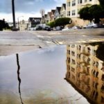 Rain puddle reflecting an urban apartment building, with swirling multicolored oil slick on the surface.