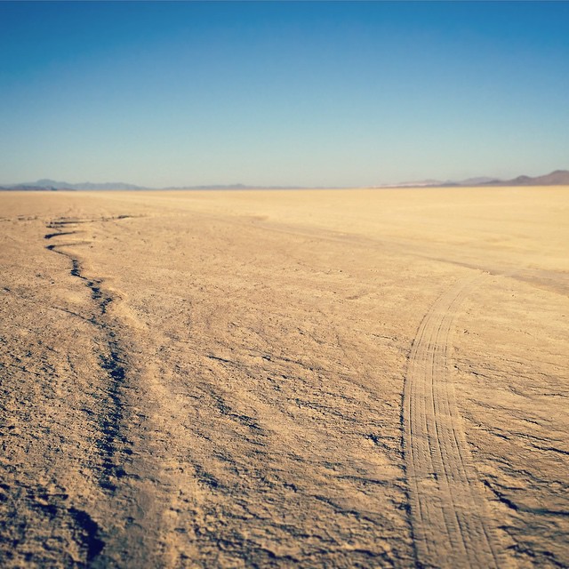 A tire track and dry stream channel run parallel toward the horizon, in a sparse desert landscape.
