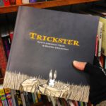 One hand holding a book with a rabbit illustration and the title "Trickster: Native American Tales, A Graphic Collection"