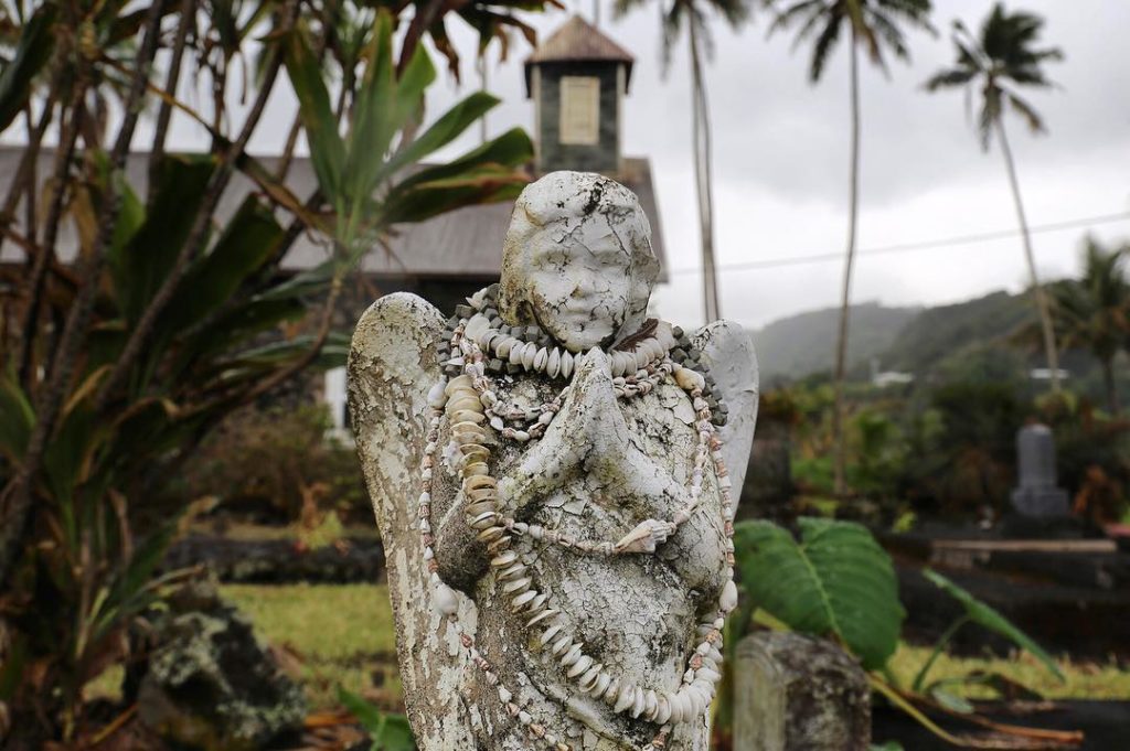Weathered angel statue in the cemetery of an old church under a cloudy sky in a tropical setting.