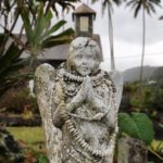 Weathered angel statue in the cemetery of an old church under a cloudy sky in a tropical setting.