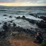 Woman crouched with camera on a rocky red-dirt shoreline, crashing waves are in the foreground and a colorful sunset with storm clouds lies in the distance.