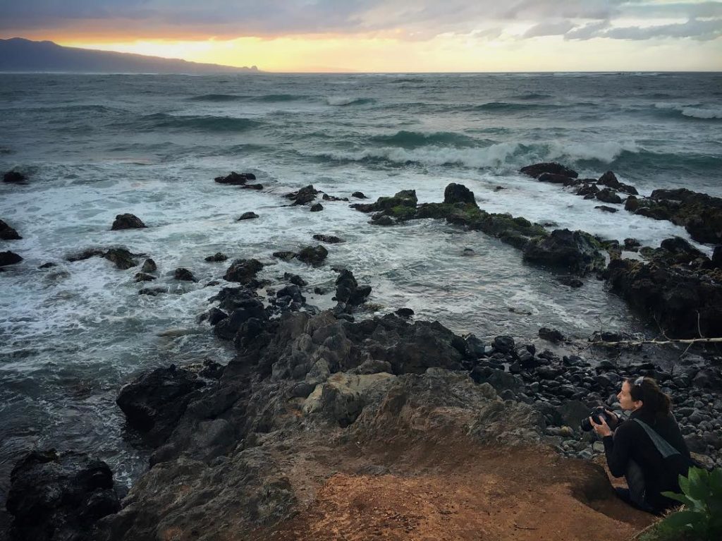 Woman crouched with camera on a rocky red-dirt shoreline, crashing waves are in the foreground and a colorful sunset with storm clouds lies in the distance.