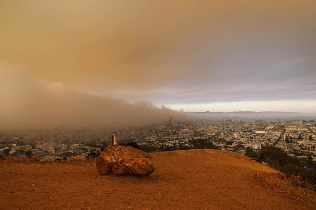 Metal water bottle sitting on a large rock, overlooking a cityscape with a heavy wall of smoke approaching from the left.