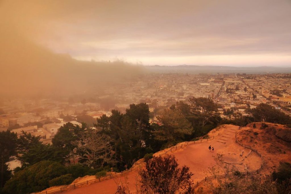 View from above a terraced city park where two people walk and heavy wildfire smoke approaches from the left.